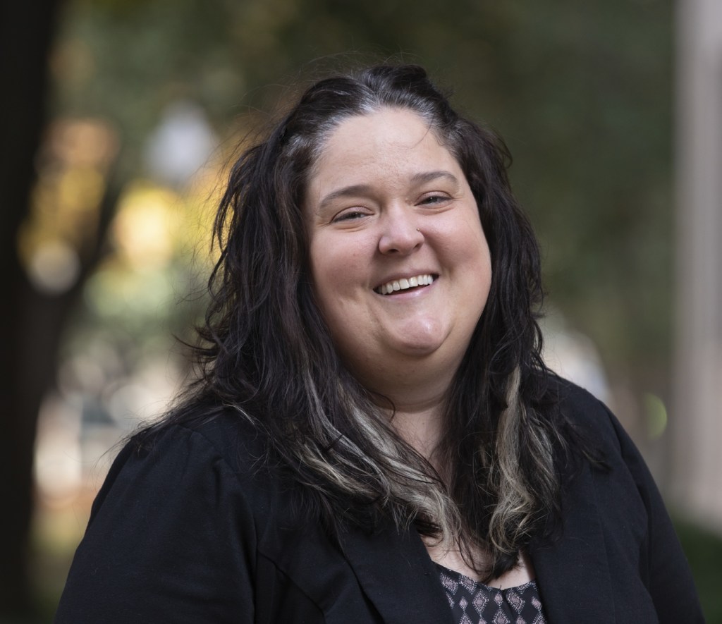 A headshot of a white female with dark hair wearing a black suit jacket with patterned top underneath and smiling. Blurry trees in the background. 